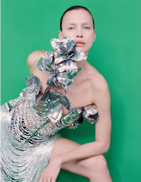 Elegant woman poses in a metallic fringed dress with flower accents against a vivid green backdrop, fashion-forward and striking.