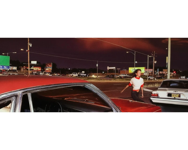 A young woman crosses a street at night near cars, streetlights, and billboards, creating a vibrant yet somewhat gritty urban scene.