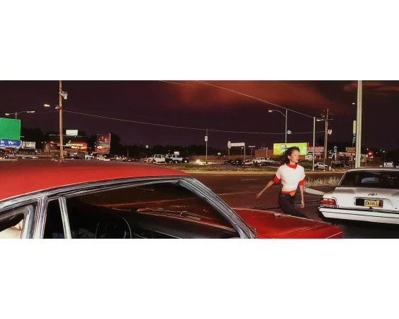 A young woman crosses a street at night near cars, streetlights, and billboards, creating a vibrant yet somewhat gritty urban scene.