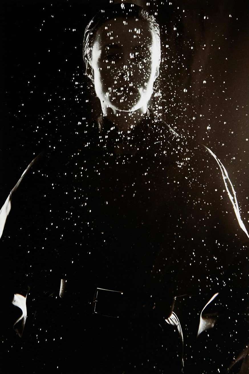 Monochrome portrait of a person in a dark jacket behind a cascading pattern of water droplets.