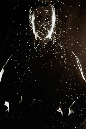 Monochrome portrait of a person in a dark jacket behind a cascading pattern of water droplets.