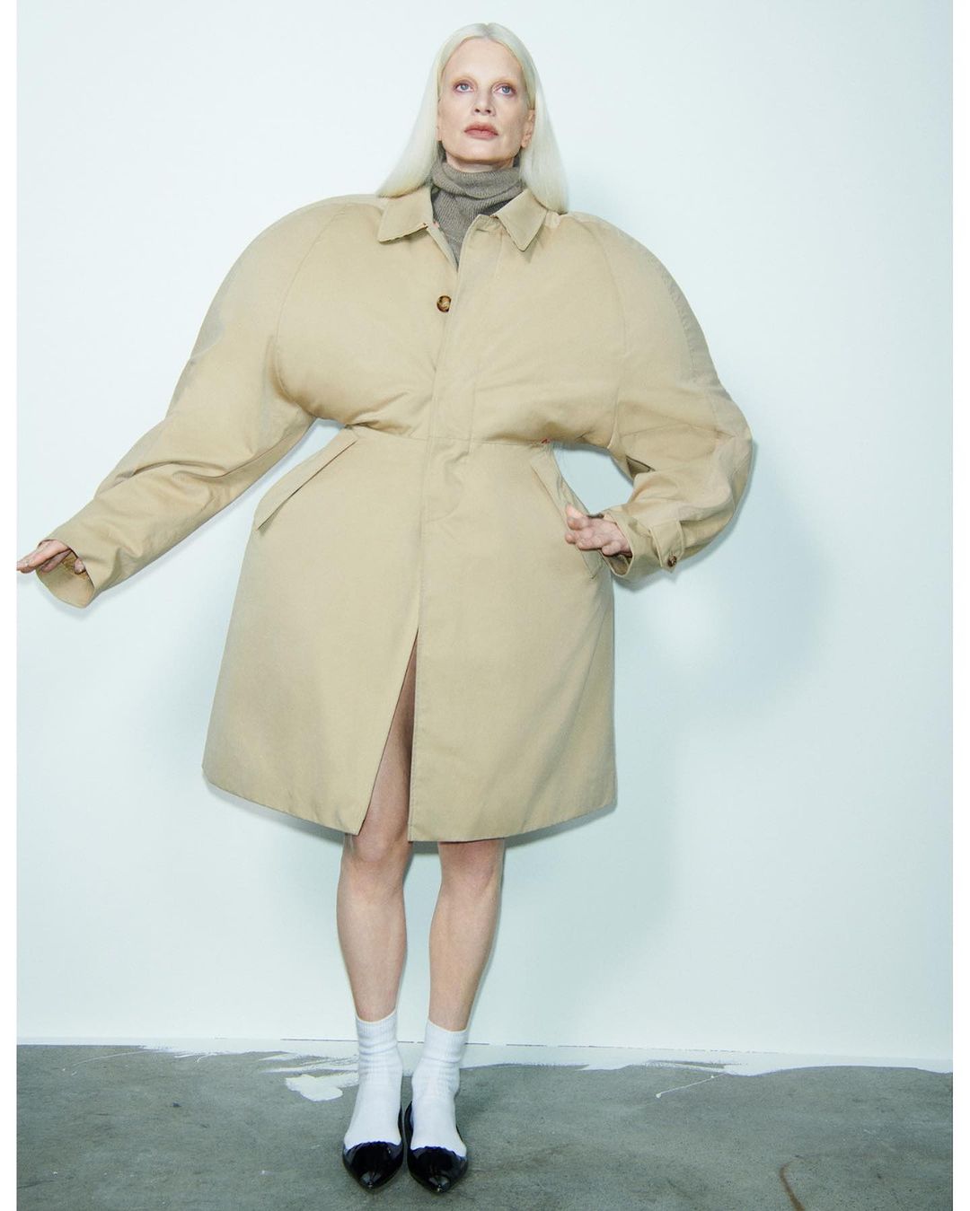 Adult female fashion model poses in an oversized tan trench coat with socks and patent leather shoes against a grey and white background.