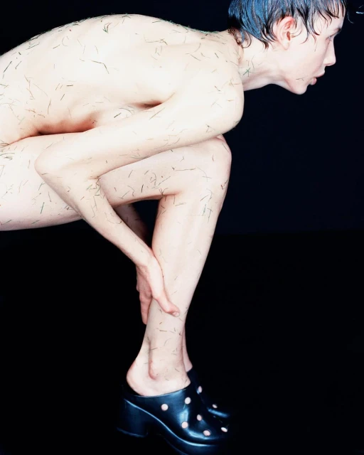 Androgynous adult poses in high-heeled clogs against a black backdrop, body art featuring tiny strands covering torso and legs.