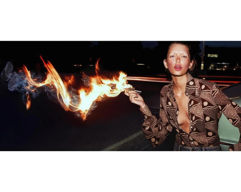 Adult woman in patterned shirt exhales a plume of smoke and flame in a nighttime outdoor setting, framed by streetlights and a dark sky.