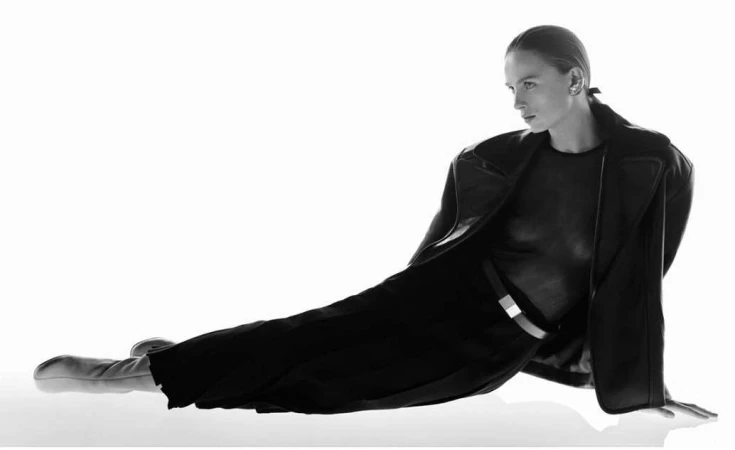 Monochrome fashion editorial portrait of a seated model wearing a dark coat and trousers against a bright white background.