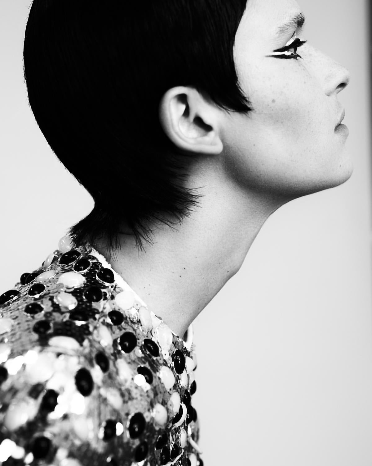 Headshot of a woman in profile wearing a sequined top and black eyeliner with short black hair against a light gray background.