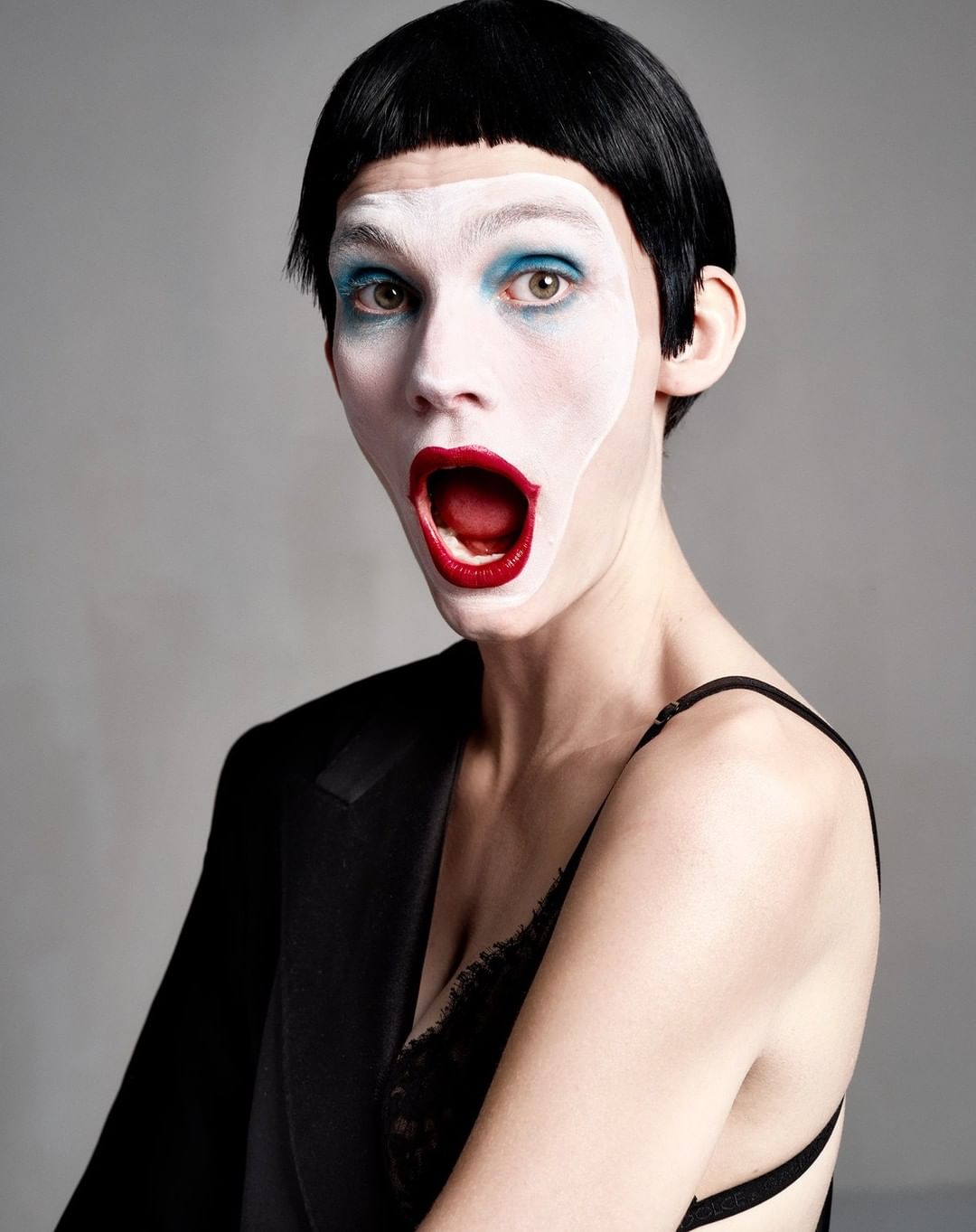 Portrait of a person with dramatic white face makeup, red lipstick, and teal eyeshadow wearing a black jacket and lace lingerie.