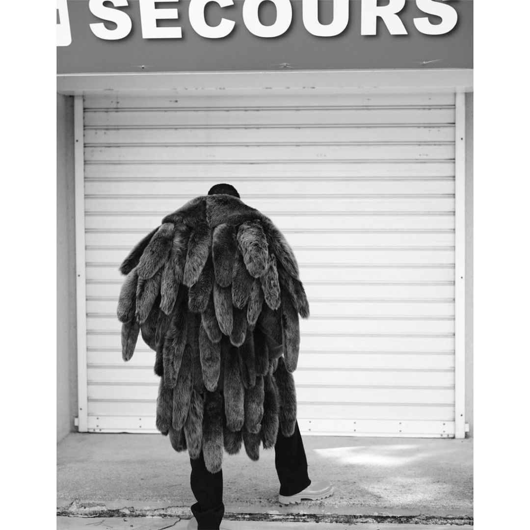 Person with their back to the camera, wearing a unique fur coat, standing outside of a building with a white metal door and a sign.