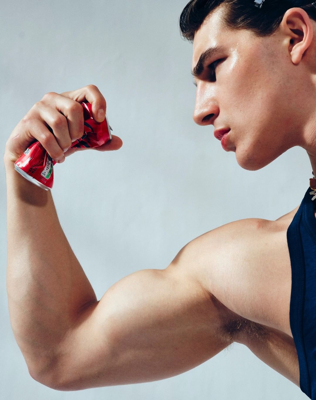 Adult male flexing bicep while crushing a red soda can in a close-up studio shot with bright lighting and light grey background.