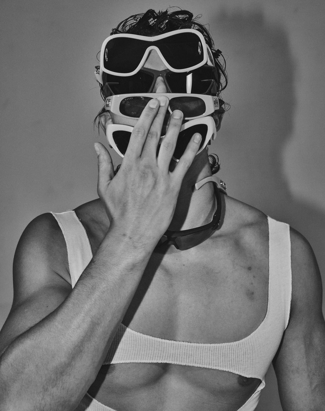 Monochrome portrait of a muscular adult male wearing a ribbed top and a stack of sunglasses, with his hand near his face.