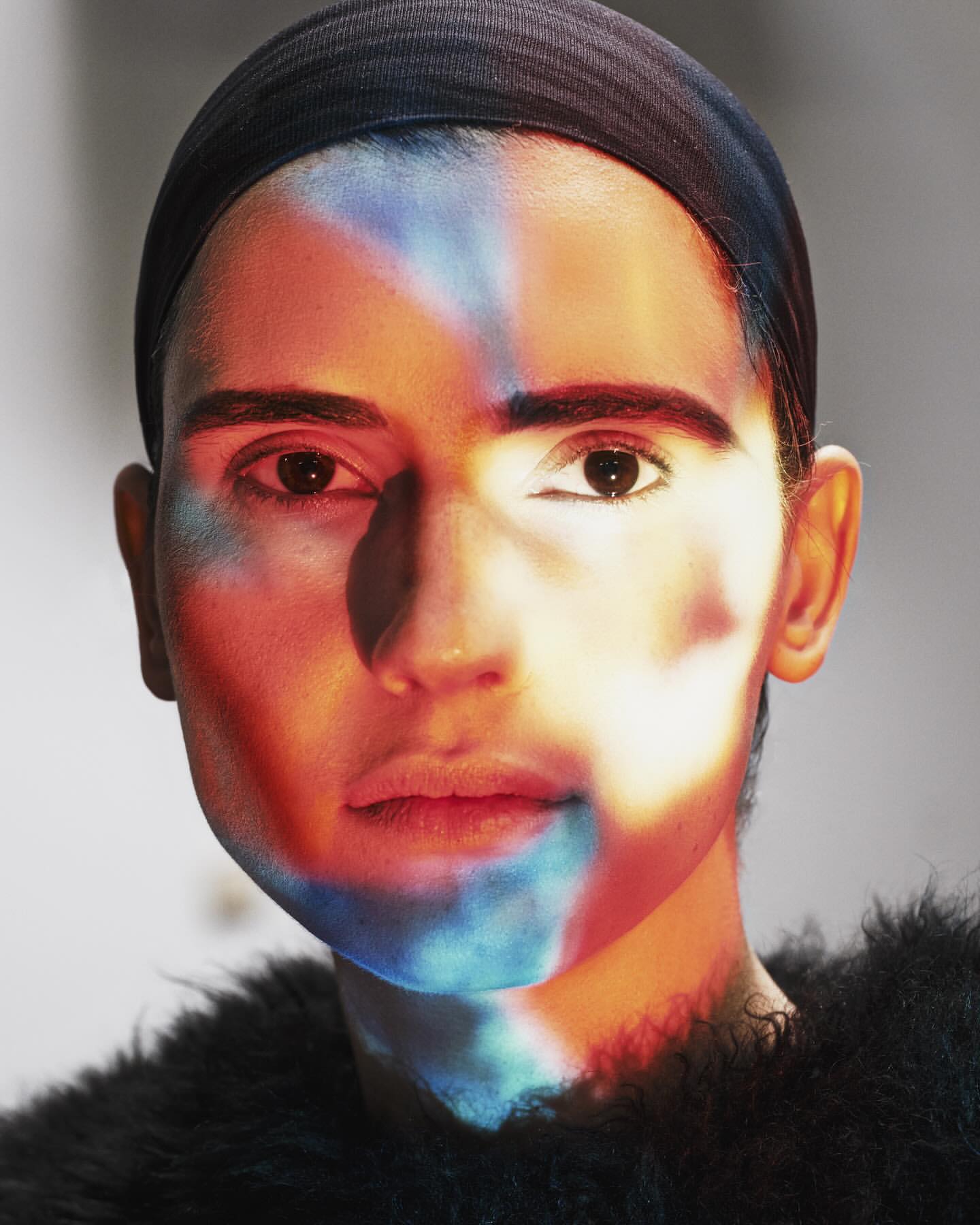 Close-up portrait of a person with short hair and a black head covering, featuring dramatic lighting with orange and blue hues on the face.