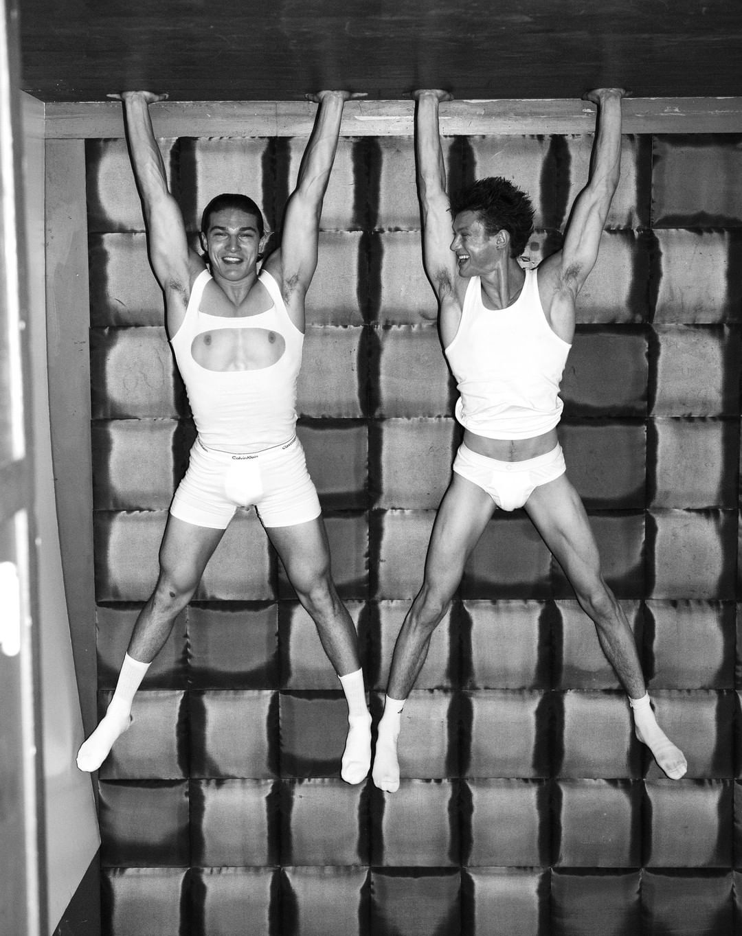 Black and white shot of two shirtless men in athletic shorts and socks hanging from an overhead bar indoors with a textured wall.