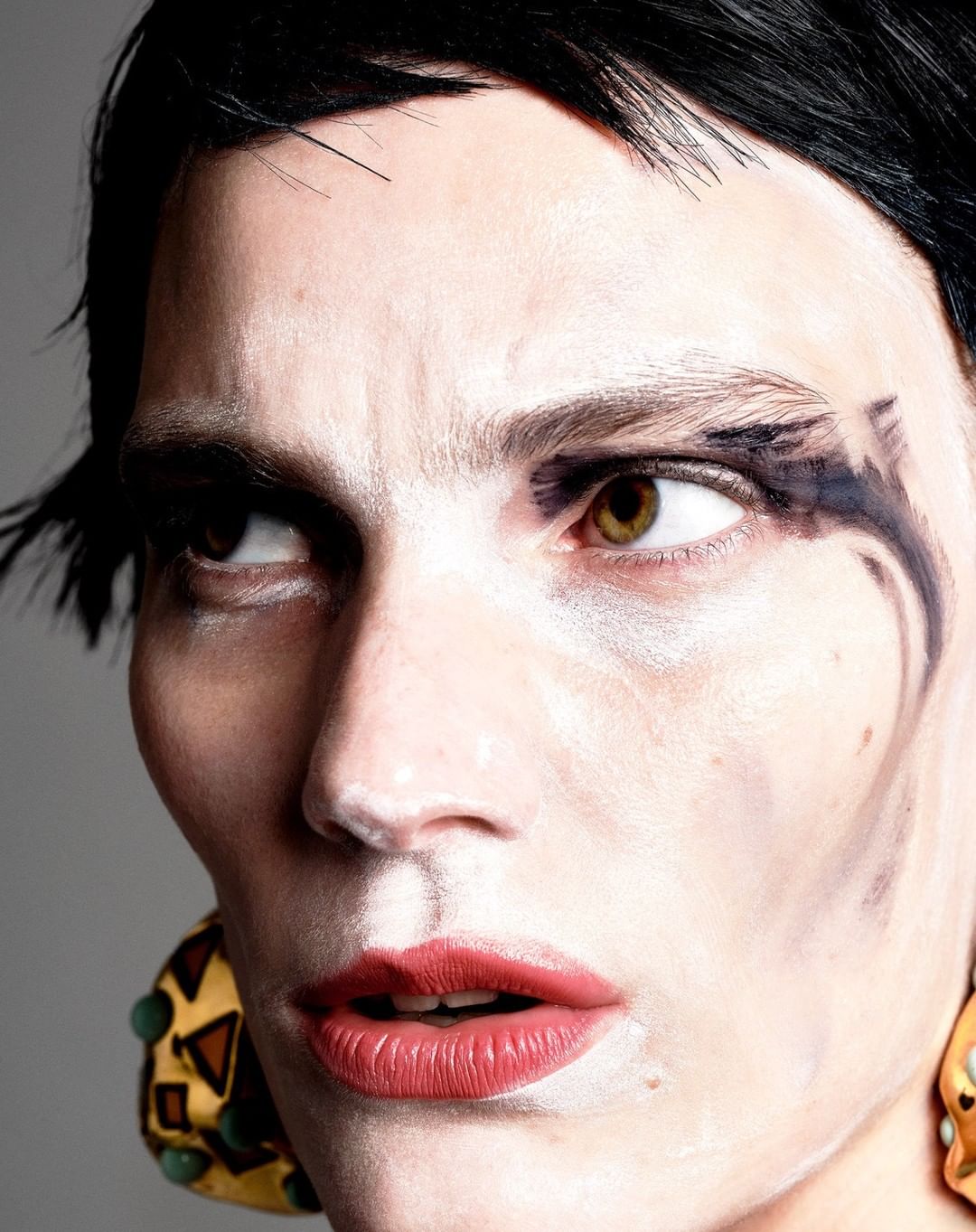 Close-up portrait of a person with black hair and striking makeup, featuring bold eye accents and red lipstick. The image highlights fashion makeup.