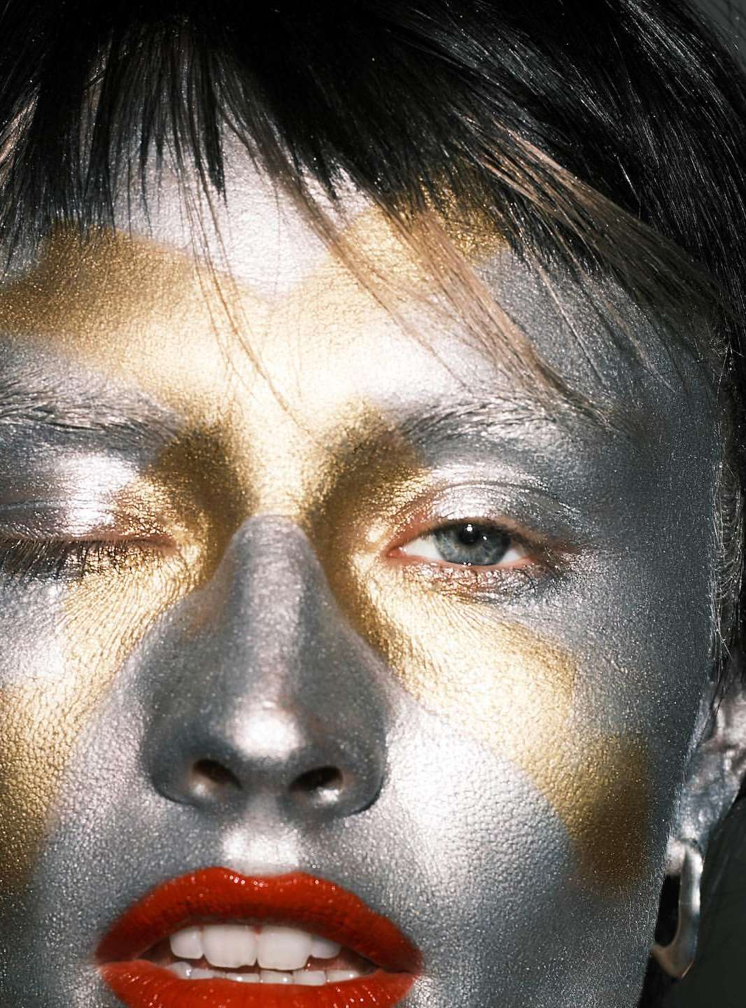 Close-up of a woman's face with silver and gold face paint, red lips, and one eye open looking at the camera.
