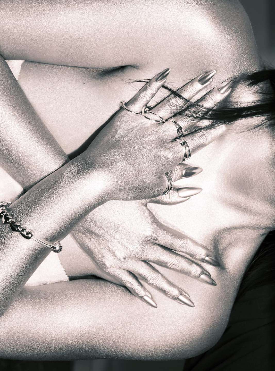 Close-up of an adult woman with metallic skin, wearing rings and a bracelet, and posing with her hands covering her chest.