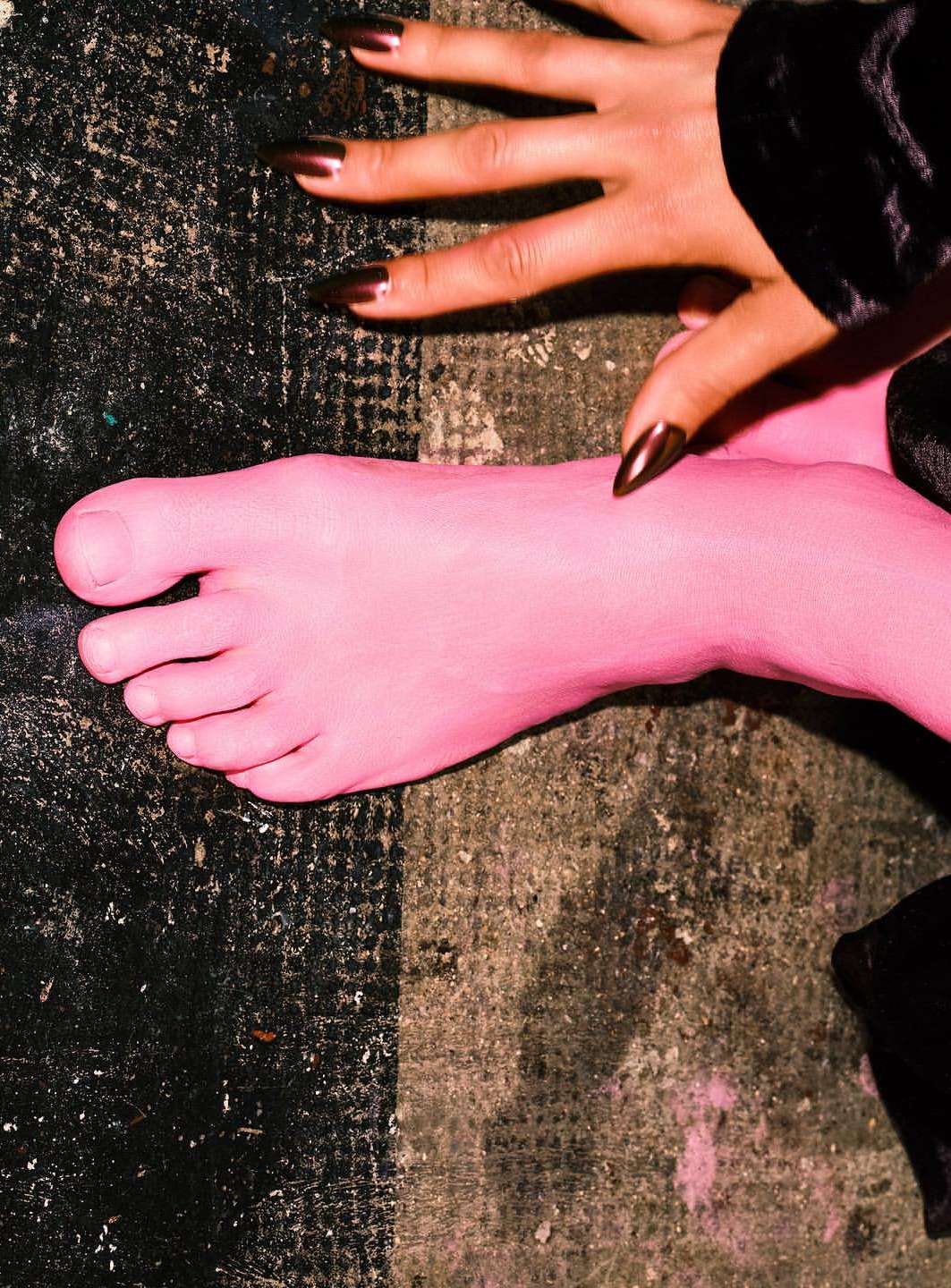 Close-up studio shot of a pink-painted foot and a hand with metallic, stiletto-shaped nails on a mottled black and gray surface.