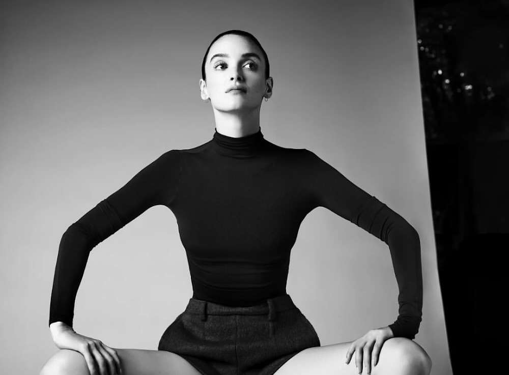 Black and white portrait of a young adult woman in dark clothing posing in a studio setting with a neutral background and soft lighting.