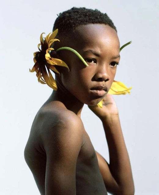 Portrait of a dark-skinned child with flowers around their head, looking sideways in a studio setting against a plain white backdrop.