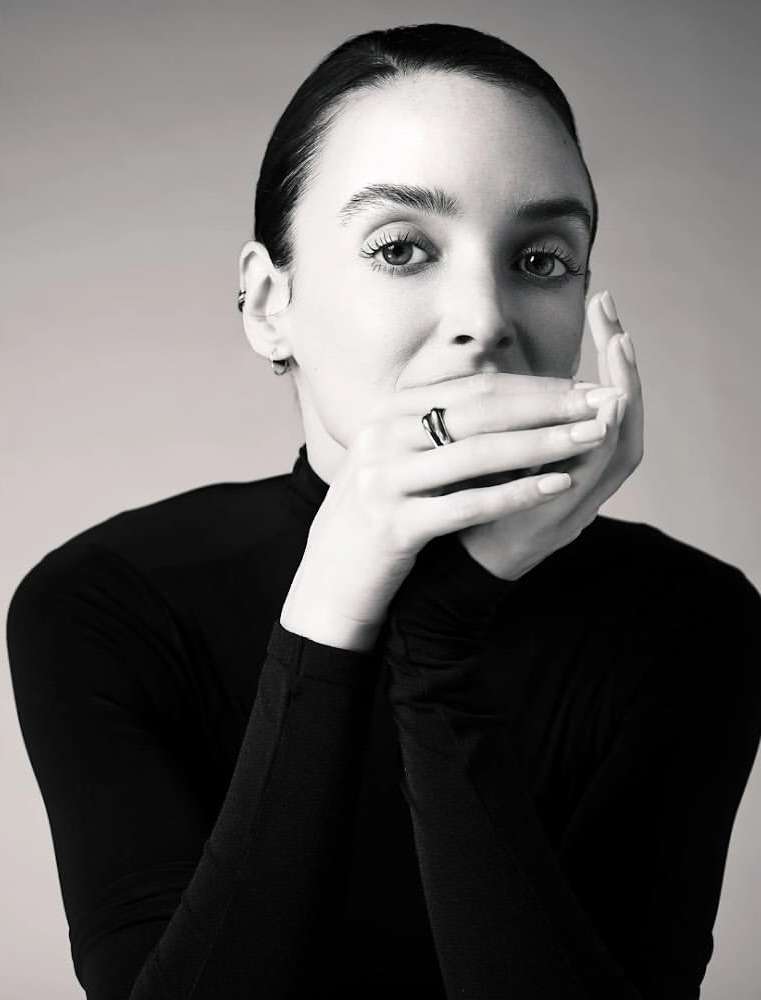 Monochrome portrait of an adult female wearing black top, face partially covered by hands, showcasing a ring against a light grey background.