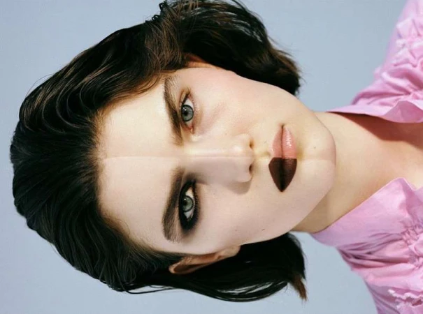 Portrait of a woman with dark hair and pink shirt, lying on her side with dramatic eye makeup and partially dark lipstick in a studio setting.