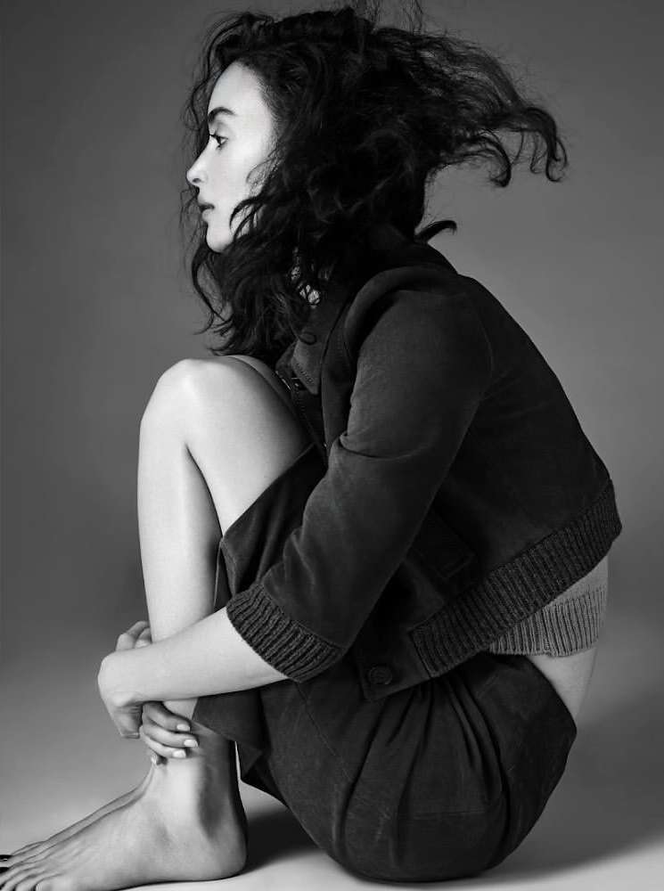 Monochrome portrait of a barefoot woman with curly hair, seated and hugging her knees, wearing a blazer and skirt against a grey background.
