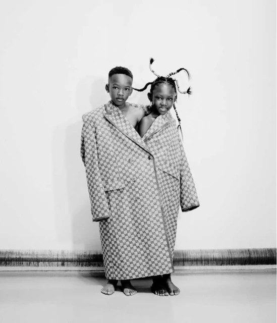 Two children share an oversized grey, patterned coat, standing barefoot against a stark white wall in an indoor studio setting.