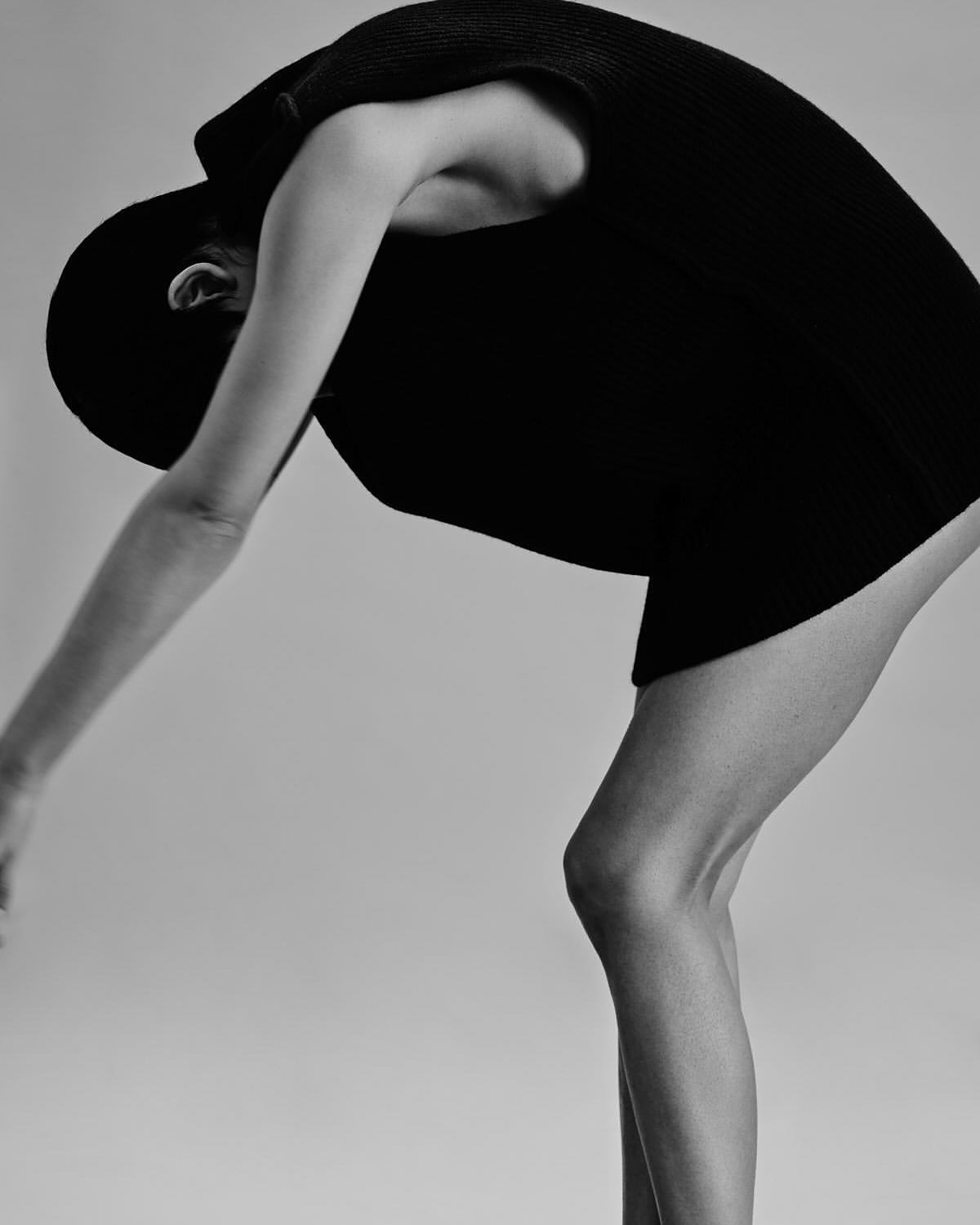 Monochrome fashion shot of a woman in a dark dress bending forward in a studio setting with a plain background, emphasizing form and texture.