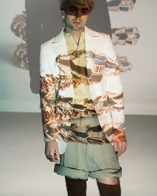 Blonde man in studio wearing a white jacket, shorts and patterned top, posing with a geometric pattern projected onto the wall and clothing.