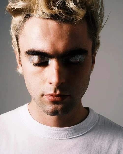 Close-up portrait of a young adult man with dyed blonde hair, wearing a white t-shirt and silver glitter makeup on his closed eyelids.