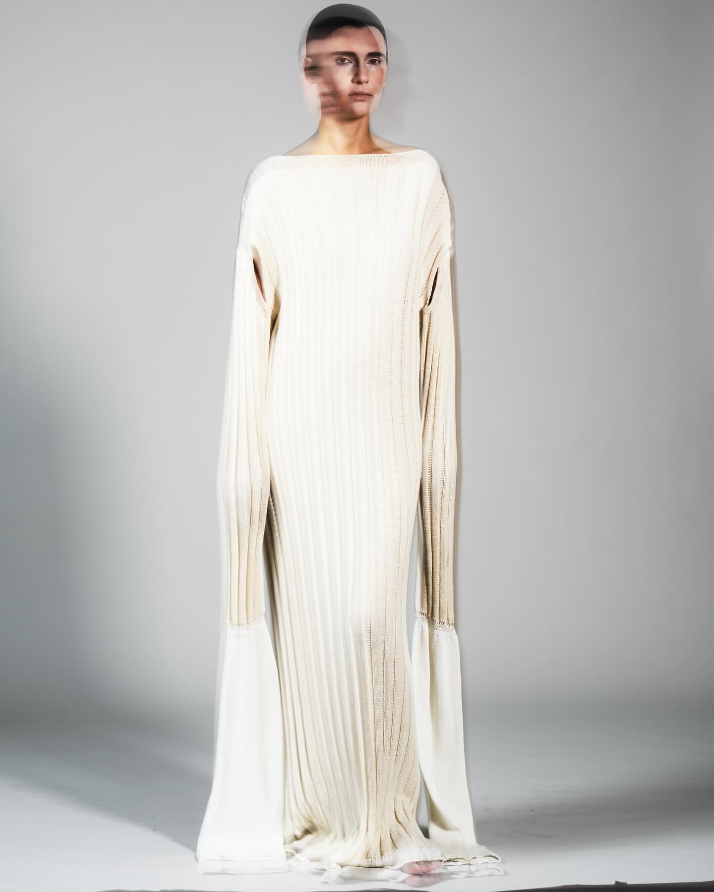 Woman poses in a full-length, ribbed, light beige dress with sleeves that extend into long, flowing, off-white fabric extensions.
