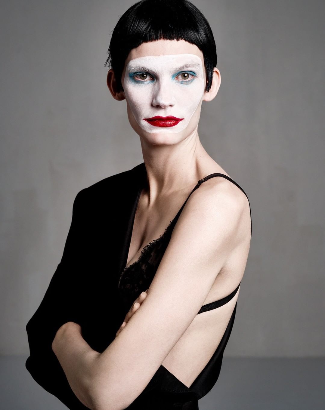 Portrait of a female model wearing white face makeup, dark lipstick, a black top, and a dark jacket, posing with her arms crossed.
