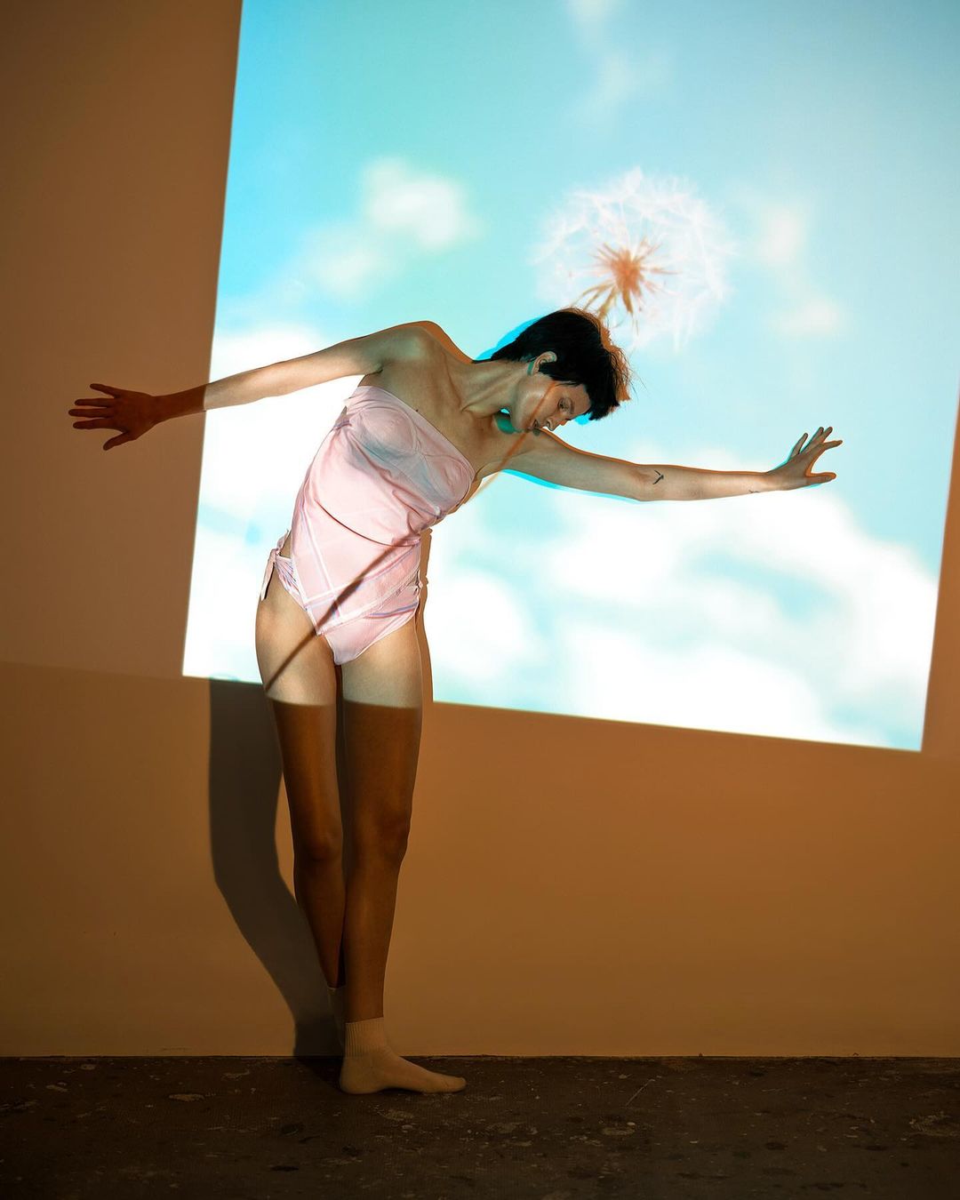 A woman strikes an artistic pose in a light pink bathing suit in front of a projection of a blue sky with a dandelion seedhead.