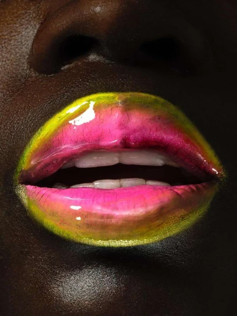 Close-up of an adult woman's face with pink and yellow glossy lipstick on full lips, visible teeth, dark skin tone, and studio lighting.