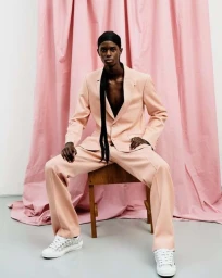 Black man seated on a wooden chair wearing a pink suit and white sneakers against a pink fabric backdrop