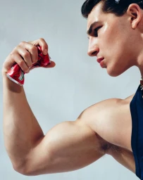 Close-up of a muscular man flexing his bicep while crushing a red soda can in his hand against a plain white background.