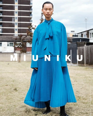 Model in vibrant blue pleated skirt and matching tie-collar blouse standing outdoors in front of a residential building.