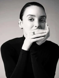 Close-up black and white portrait of a woman in a black turtleneck holding her hands up to cover her lower face.