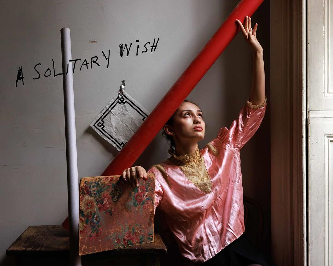 Young woman poses indoors in a pink top with red and gray cylindrical objects. Floral artwork and wall text add to the scene.