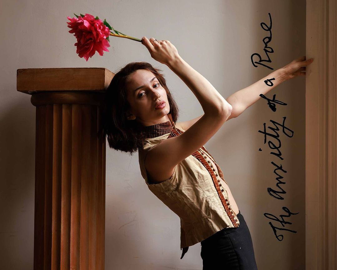 A young woman in a vintage top poses with a large red flower near a pillar, indoors with soft natural light and handwritten text overlay.