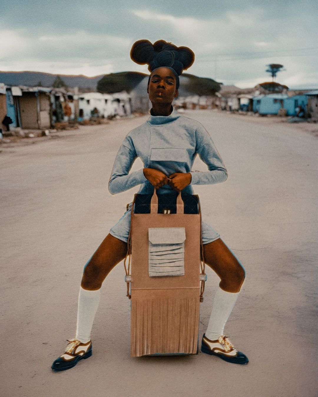 Stunning fashion shot features a black model in light-blue outfit with a geometric bag-like contraption, posing on a remote desert road.