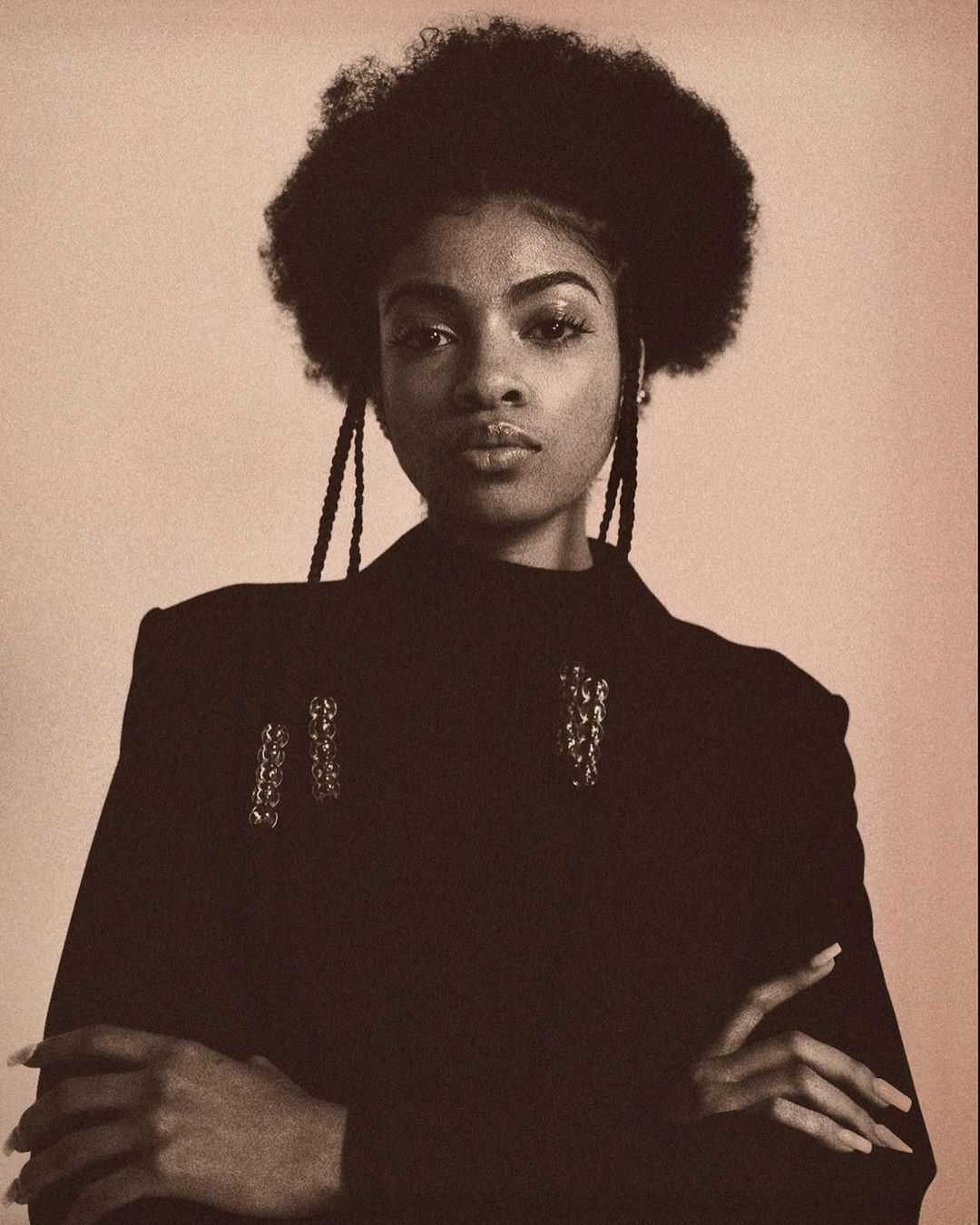 Portrait of an adult woman with afro hairstyle, wearing black jacket with ornamental details, posing with arms crossed against light background.