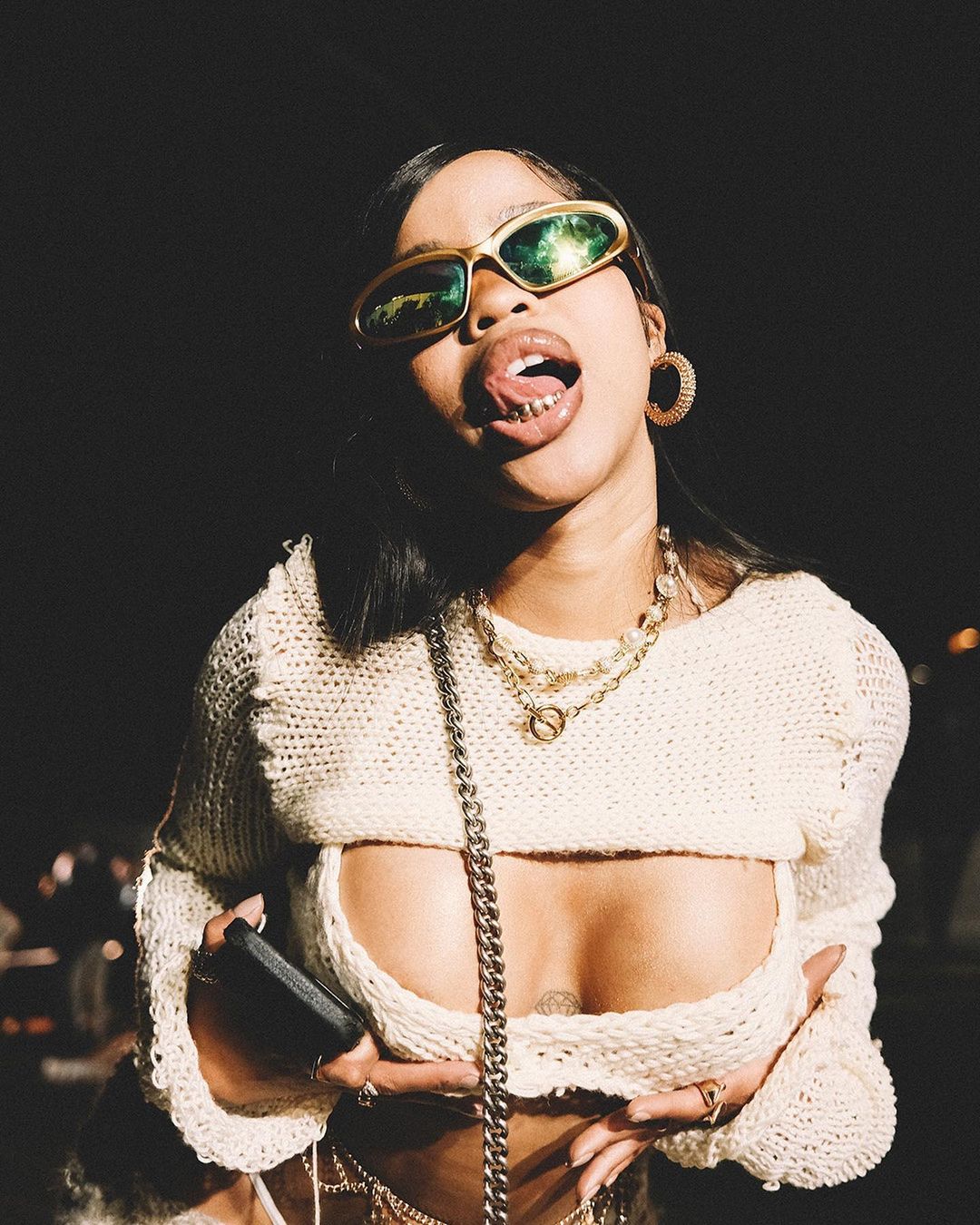 Portrait of an African American woman wearing retro sunglasses, a white sweater, hoop earrings, and layered necklaces against a dark background.
