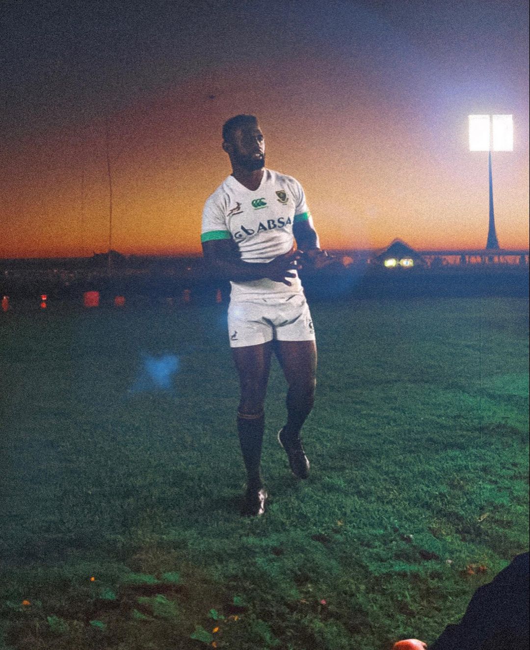 Rugby player in uniform stands on the field with stadium lights in the background and a golden sky at sunset or sunrise.
