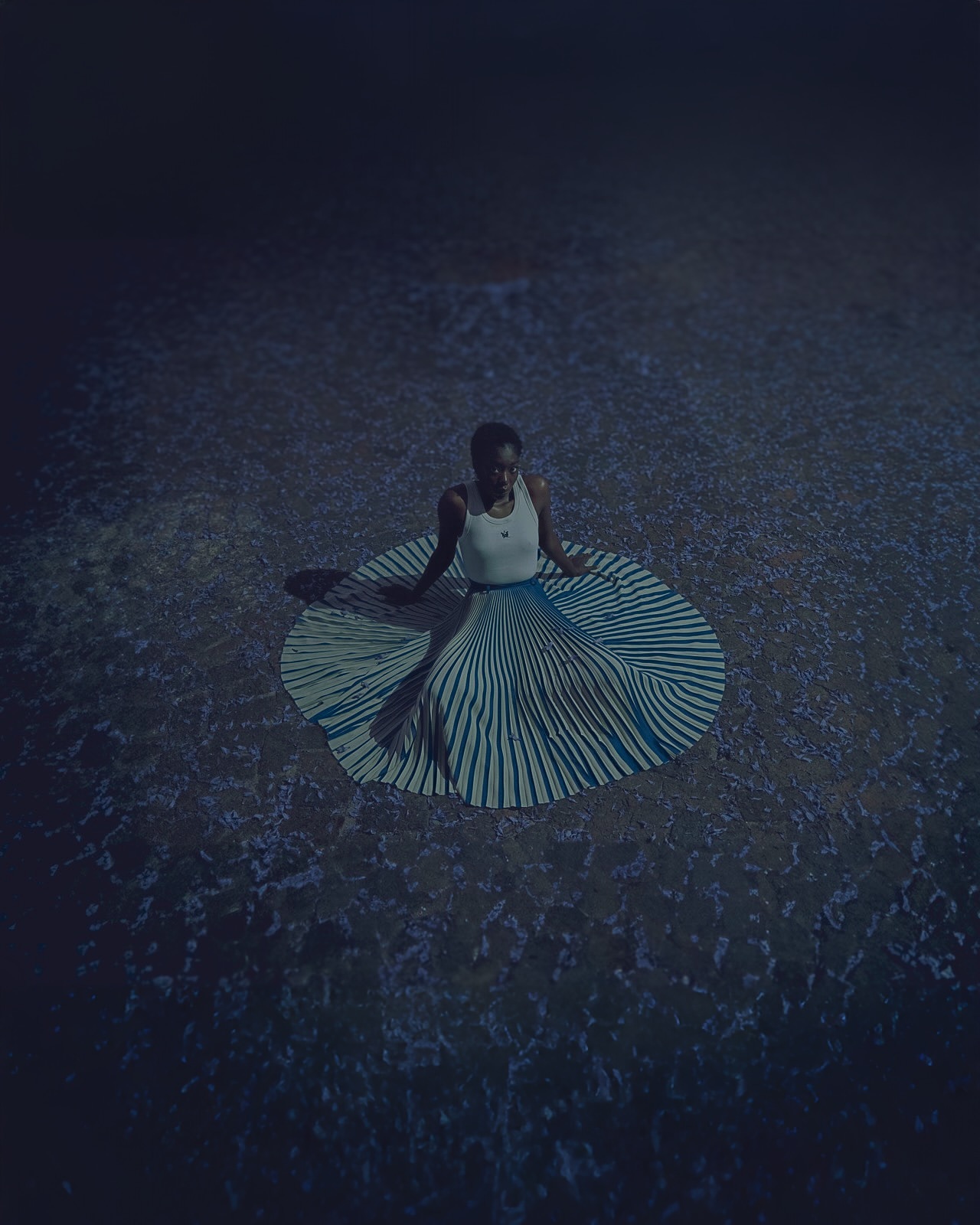Woman seated in striped skirt on textured ground in a dim, moody scene with a high-angle view, creating a captivating and artistic portrait.