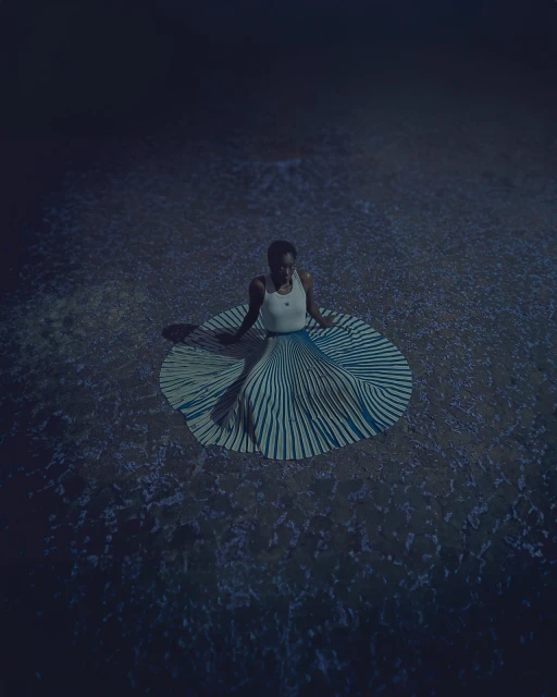 Woman seated in striped skirt on textured ground in a dim, moody scene with a high-angle view, creating a captivating and artistic portrait.