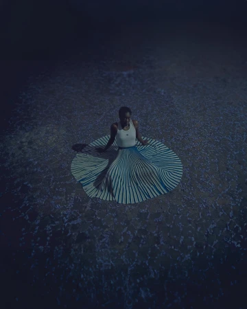Woman seated in striped skirt on textured ground in a dim, moody scene with a high-angle view, creating a captivating and artistic portrait.