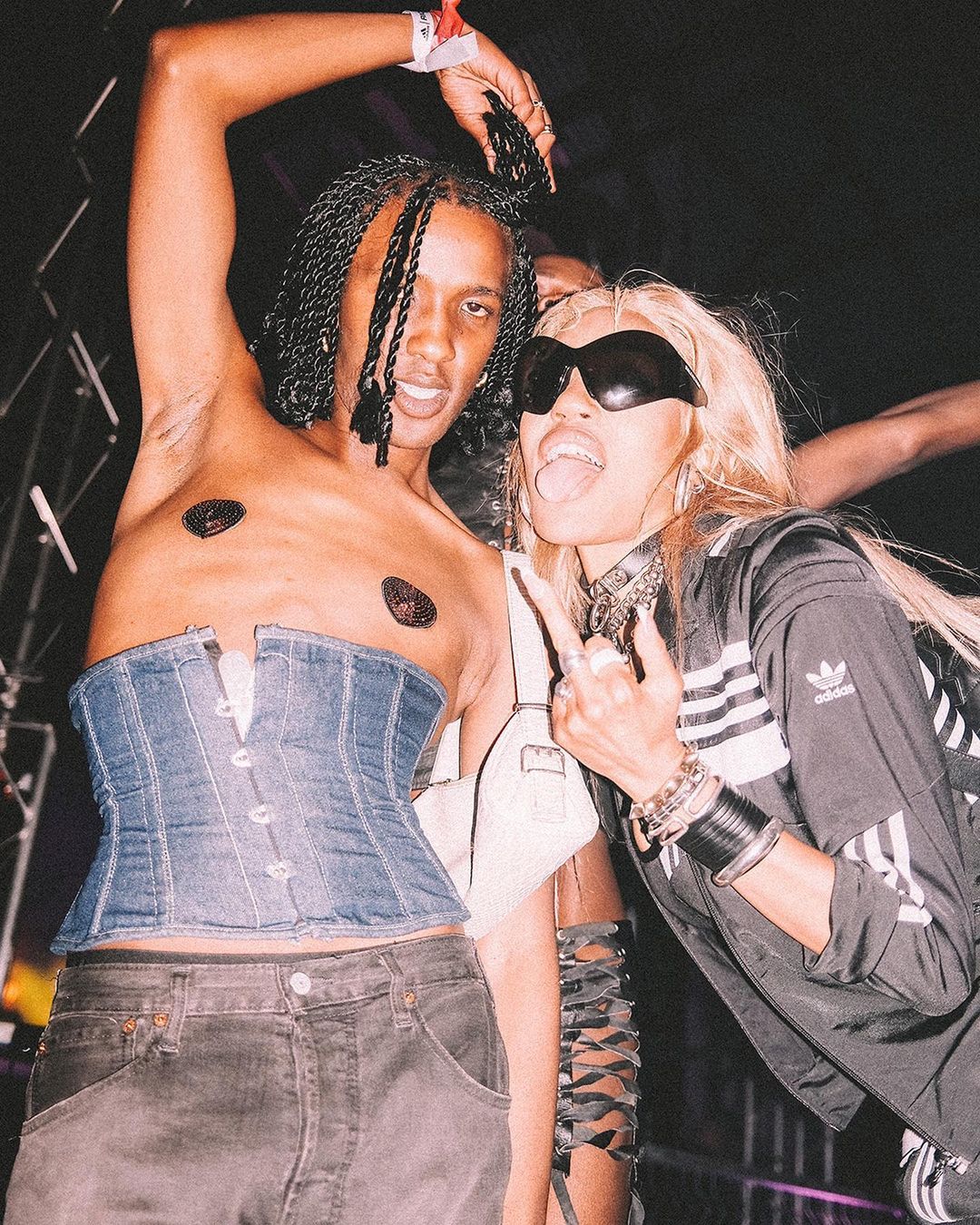 Two young adults pose together indoors at a party with dark lighting. One is wearing a denim corset, and the other is wearing sunglasses.