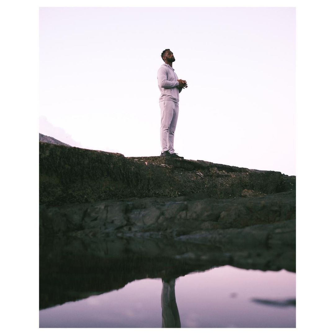 A man in a grey sweatsuit stands on a rock formation near calm water, looking upwards with bright sky in the background.