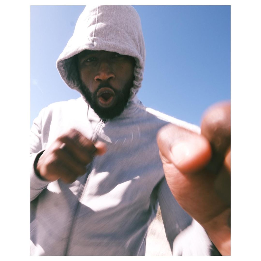 A man with a beard and hoodie, looking directly at the camera with an open mouth, is standing outside against a clear blue sky.