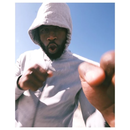 A man with a beard and hoodie, looking directly at the camera with an open mouth, is standing outside against a clear blue sky.